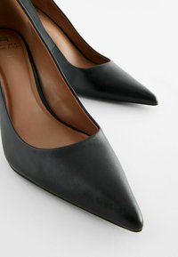 Tacones altos de cuero negro con punta afilada, textura suave y diseño minimalista; cuentan con un forro interior en tono marrón.