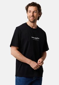 Zwarte katoenen t-shirt met korte mouwen, met een witte geprinte logo op de voorkant. Draagt met blauwe jeans. Eenvoudig, casual ontwerp.