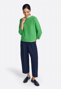 Groene, gestructureerde trui met een witte kraag, gecombineerd met losse marineblauwe broek en zwarte instappers. Model glimlacht en raakt de kraag aan.