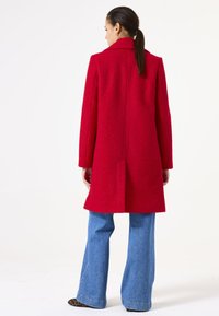 Manteau en laine rouge avec une finition texturée, coupe droite et fente au dos. Associé à un jean bleu à jambes larges.