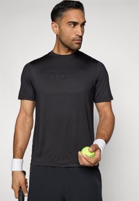 Juodas trumpas rankovėmis sportinis marškinėlis su subtilia iškilminga logotipo imitacija, jį dėvi vyras, laikantis žalią teniso kamuoliuką, su baltais riešų raiščiais.