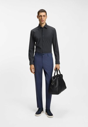 Uomo che indossa una camicia nera a maniche lunghe con motivo, pantaloni blu, scarpe da ginnastica blu, che tiene una grande borsa di pelle nera su uno sfondo bianco.