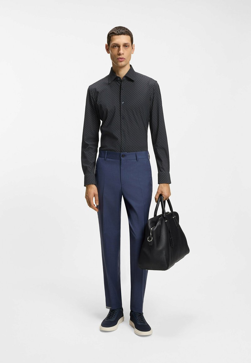 Homme portant une chemise noire à motifs à manches longues, un pantalon bleu, des baskets bleues, tenant un grand sac à main en cuir noir sur un fond blanc.