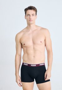 Schwarze Boxershorts mit einem bordeauxfarbenen Bund, der das "PUMA"-Logo in Weiß trägt. Glattes Material, Oberschenkel-lange, figurbetonte Passform.