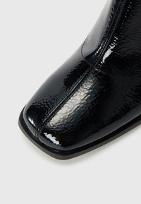 Chaussure en cuir verni noir avec un bout carré et des coutures centrales visibles sur fond blanc.