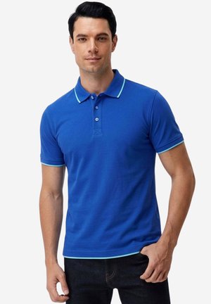 Uomo con polo blu brillante a maniche corte con bordo azzurro chiaro sul colletto e sulle maniche, in piedi con una mano in tasca.