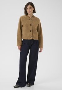 Žena oblečená v hnedom vlnenom sáčku na gombíky s dlhými rukávmi, tmavých širokých nohaviciach a čiernych topánkach so cvokmi, stojaca na bielom pozadí.