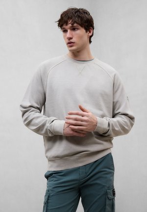 Giovane uomo con capelli ricci scuri che indossa una felpa beige e pantaloni cargo turchesi, in piedi contro uno sfondo grigio chiaro uniforme, con le mani incrociate.