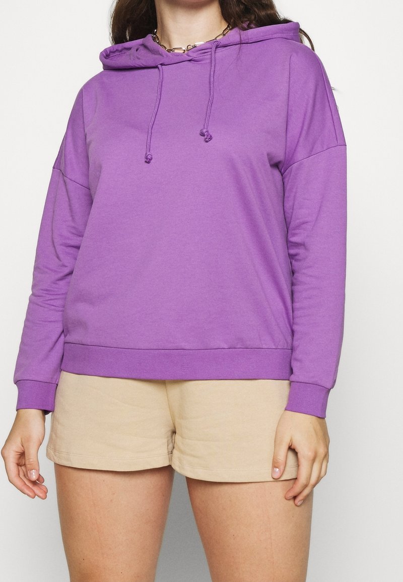 Personne portant un sweat à capuche violet avec cordons et un short beige, debout devant un fond clair uni.