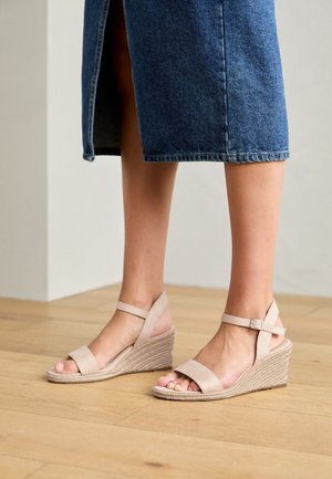 Ben iført en knælængde denimnederdel og beige kile-sandaler med anklerem, stående på et trægulv.
