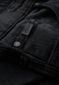 Mustapari denim-kangas, jossa on teksturoitu pinta, metallinappi ja ommeltu etiketti, jossa lukee "one more daps".