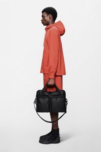 Svart strukturert veske med to frontlommer, korte håndtak og en avtagbar skulderstropp, holdt av en person iført et korallfarget antrekk.