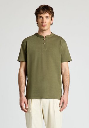 Junger Mann trägt ein olivgrünes Henley-Shirt mit kurzen Ärmeln und lockere cremefarbene Hosen, steht vor einem schlichten hellen Hintergrund.