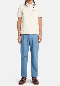 Kremowa koszulka polo z zapięciem na guziki przy kołnierzyku i kieszenią na piersi, zestawiona z jasnoniebieskimi spodniami o prostych nogawkach oraz brązowymi skórzanymi butami.