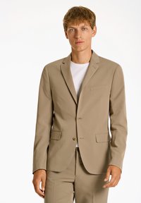 Beige Sakko mit zwei Knöpfen, Reverskragen und einer Brusttasche. Getragen über einem weißen T-Shirt mit glatter Textur.