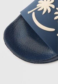 Tamsiai mėlyna šlepetė su tekstūruota padais ir matinėmis juostelėmis, kuriose yra iškilusi palmės medžio dizaino ir kreivos smėlio spalvos detalės.