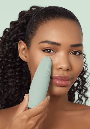 Kvinde med krøllet hår bruger en lysegrøn ansigtsrensningsenhed på sin kind mod en blød pastelbaggrund.