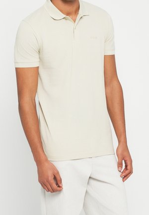 Männliches Model trägt ein schlichtes hellbeiges kurzärmeliges Poloshirt und weiße Hose vor weißem Hintergrund.
