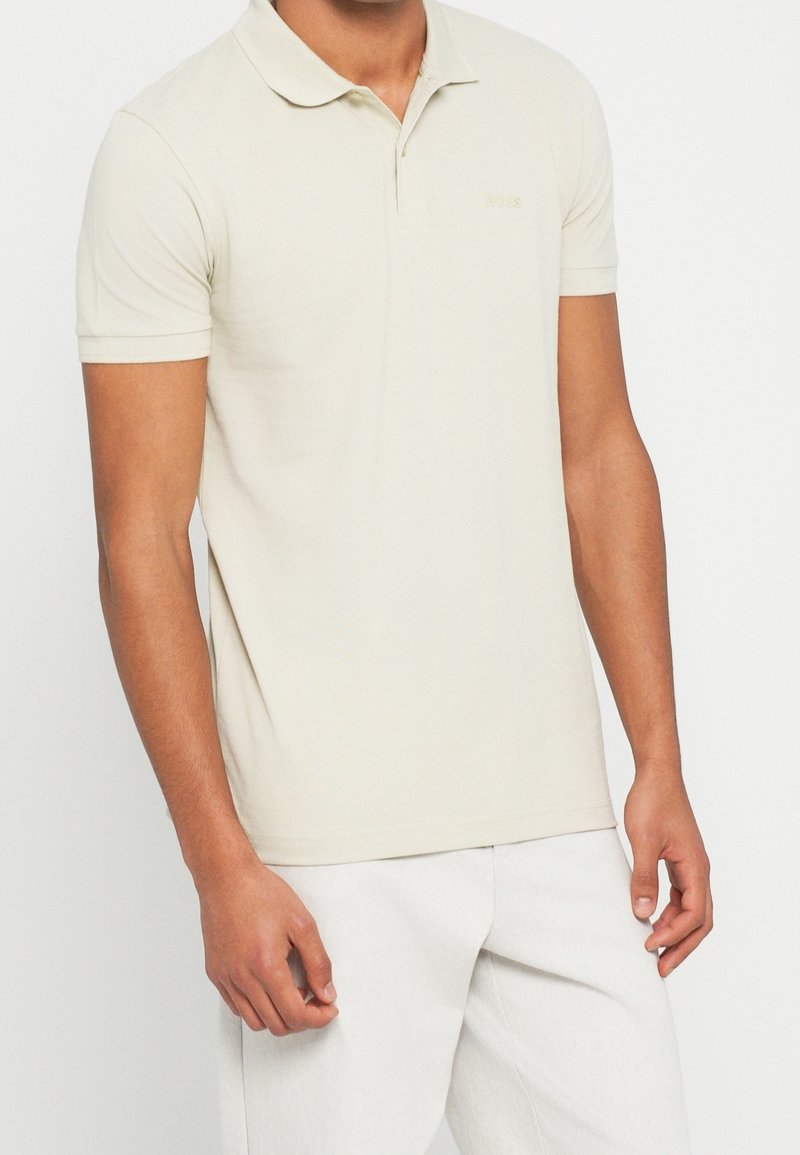 Modèle masculin portant un polo uni beige clair à manches courtes et un pantalon blanc sur un fond blanc uni.
