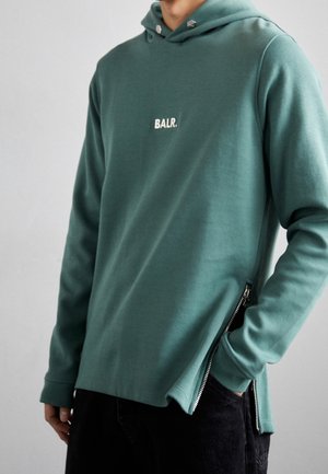 Hombre vestido con sudadera con capucha BALR de color verde azulado con logo en el frente, mangas largas y cremallera lateral parcialmente abierta, con la mano en el bolsillo, sobre un fondo liso.