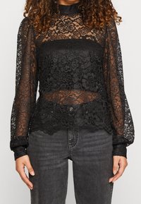 Haut noir en dentelle à manches longues avec motifs floraux, encolure à volants et accents transparents, associé à un jean foncé.