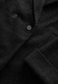 Gros plan d'un tissu de manteau en laine noire texturée avec deux boutons noirs visibles et une manche repliée.