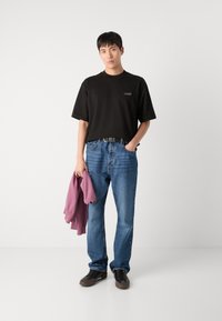 Tricou negru oversized cu mânecă scurtă, blugi denim albaștri, ținând un hanorac roz. Pantofi negri cu tălpi brune. Design simplu și croială confortabilă.