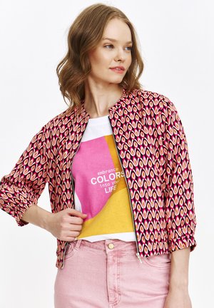 Czerwono-różowa kurtka we wzory z zamkiem z przodu, okrągłym dekoltem i marszczonymi rękawami, noszona na wielokolorowym t-shircie z grafiką.