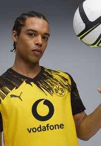 Joven atleta masculino con camiseta de fútbol amarilla y negra del Borussia Dortmund, equilibrando un balón de fútbol blanco y negro de Nike cerca de su cabeza.