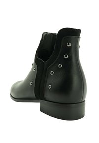 Botín de piel negra con tachuelas plateadas y tacón bajo, mostrado desde un ángulo lateral trasero sobre fondo blanco.