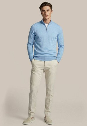 Man draagt een lichtblauwe trui met kwartrits, wit ondershirt, beige broek en beige instappers, staat met handen in de zakken.