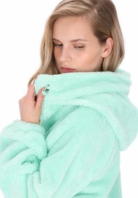 Mentaszínű plüss kapucnis pulóver puha, bolyhos textúrával, nagy kapucnival és állítható húzózsinórral. Laza szabású, mandzsettás ujjakkal.