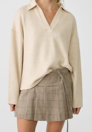 Pull beige à manches longues avec col en V, associé à une jupe plissée à carreaux avec un détail de nœud sur le côté.