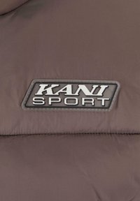 Smeđa puffer jakna s ušivenim logotipom 'KANI SPORT' u bijeloj boji na crnoj pozadini, isti�čući glatku, teksturiranu tkaninu.
