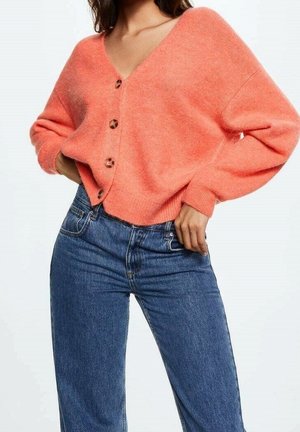 Kvinde iført løs koralfarvet cardigan med knapper og højtaljede blå jeans, hænderne hvilende på hofterne mod ensfarvet baggrund.