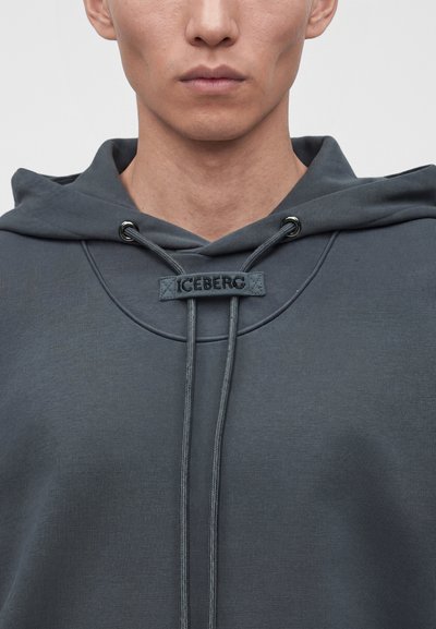 Gros plan d’une personne portant un sweat à capuche gris foncé avec « ICEBERG » brodé sur la partie avant des cordons.