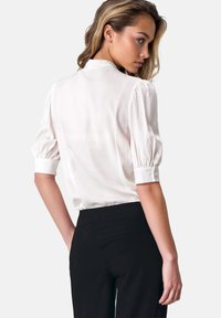 Kobieta z blond włosami, ubrana w białą bluzkę z bufiastymi rękawami i czarne spodnie z wysokim stanem, patrząca przez ramię na neutralnym tle.