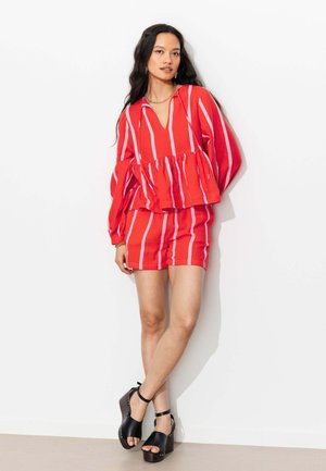 Femme portant un chemisier à manches longues rayé verticalement rouge et rose et un short assorti, debout avec les jambes croisées et des sandales compensées noires.