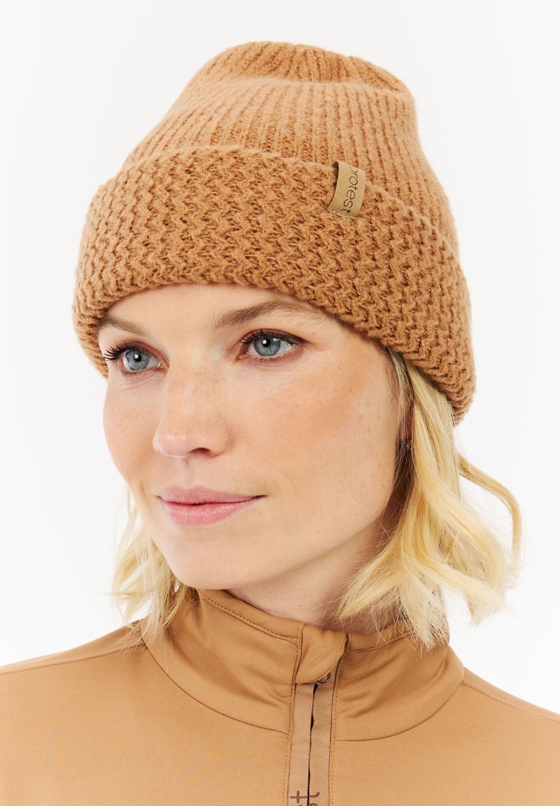 Austas megztas būgnas šiltai rudu atspalviu su rauktu viršumi ir žuvies kaulų raštu. Su mažu šviesiai rudos spalvos ženklu su logotipu šone.