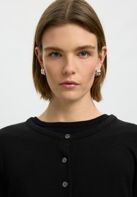 Femme aux cheveux bruns courts, portant des boucles d'oreilles créoles argentées et un haut noir boutonné sur un fond clair uni.