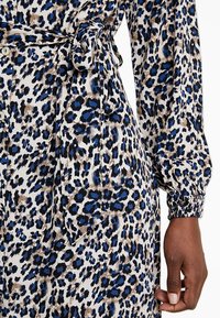 Närbild av en leopardmönstrad klänning i blått, svart och brunt med en knytning i midjan och elastiska ärmmuddar, bärs av en person med mörk hy.