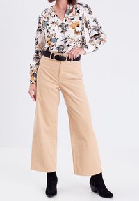 Blouse blanche à fleurs avec manches longues, col en V, et motif détaillé ; pantalon en velours côtelé beige à jambes larges ; ceinture noire ; bottines noires.