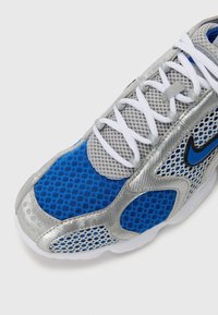 Chaussure de sport bleue et grise avec des lacets blancs, un tissu en mesh, des surcouches argentées et un logo Nike noir et bleu sur le côté.