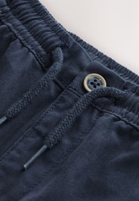 Tamsiai mėlynos spalvos elastinga liemens juosta su pintais raišteliais ir apvaliu mygtuku su keturiomis skylutėmis bei mažais įrėžtais užrašais.