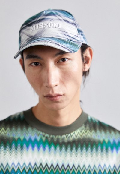 Το καπέλο διαθέτει ένα κυματιστό μοτίβο σε μπλε, πράσινο και λευκό με την επιγραφή "MISSONI" κεντημένη. Το μοντέλο φοράει ένα πράσινο πουκάμισο με ζιγκ-ζαγκ σχέδιο και στρογγυλή λαιμόκοψη.