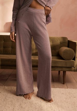 Vrouw draagt mauve geribbelde wijdvallende broek en bijpassende top, staat blootsvoets op een licht tapijt voor een olijfgroene bank.