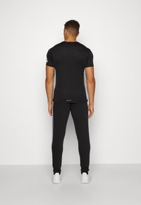 Man som står vänd mot väggen iförd svart kortärmad skjorta, svarta byxor med vita ränder och vita sneakers.