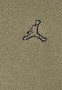 Tela de color verde oliva con una superficie texturizada que presenta un logo Jumpman bordado en negro, posicionado de forma central en la prenda.