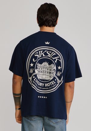 Muž v tmavomodrom tričku s bielou grafikou luxusného hotela a textom "SikSilk Luxury Hotel" na zadnej strane, v kombinácii so svetlomodrými džínsami.