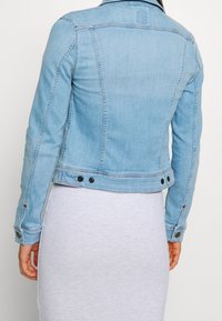 Woman wearing a light blue denim jacket over a white ribbed dress, shown from the back against a plain background.